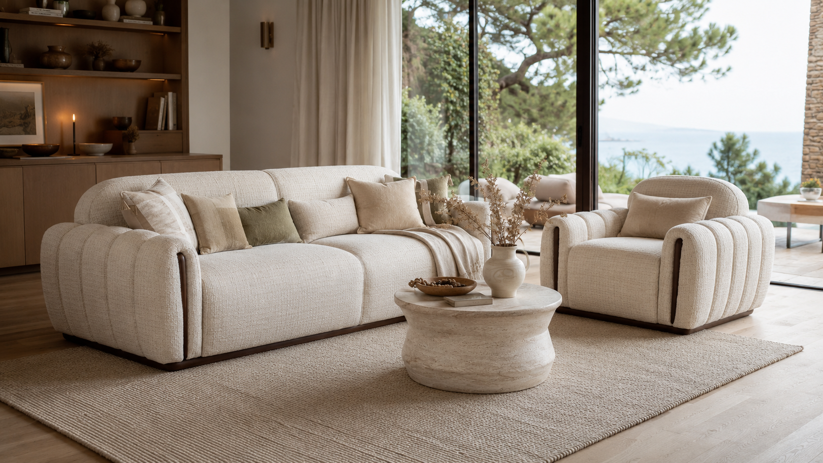 A cream-colored tufted sofa and matching armchair in a modern, sunlit living room with large windows overlooking water, a round stone coffee table, and a textured rug.