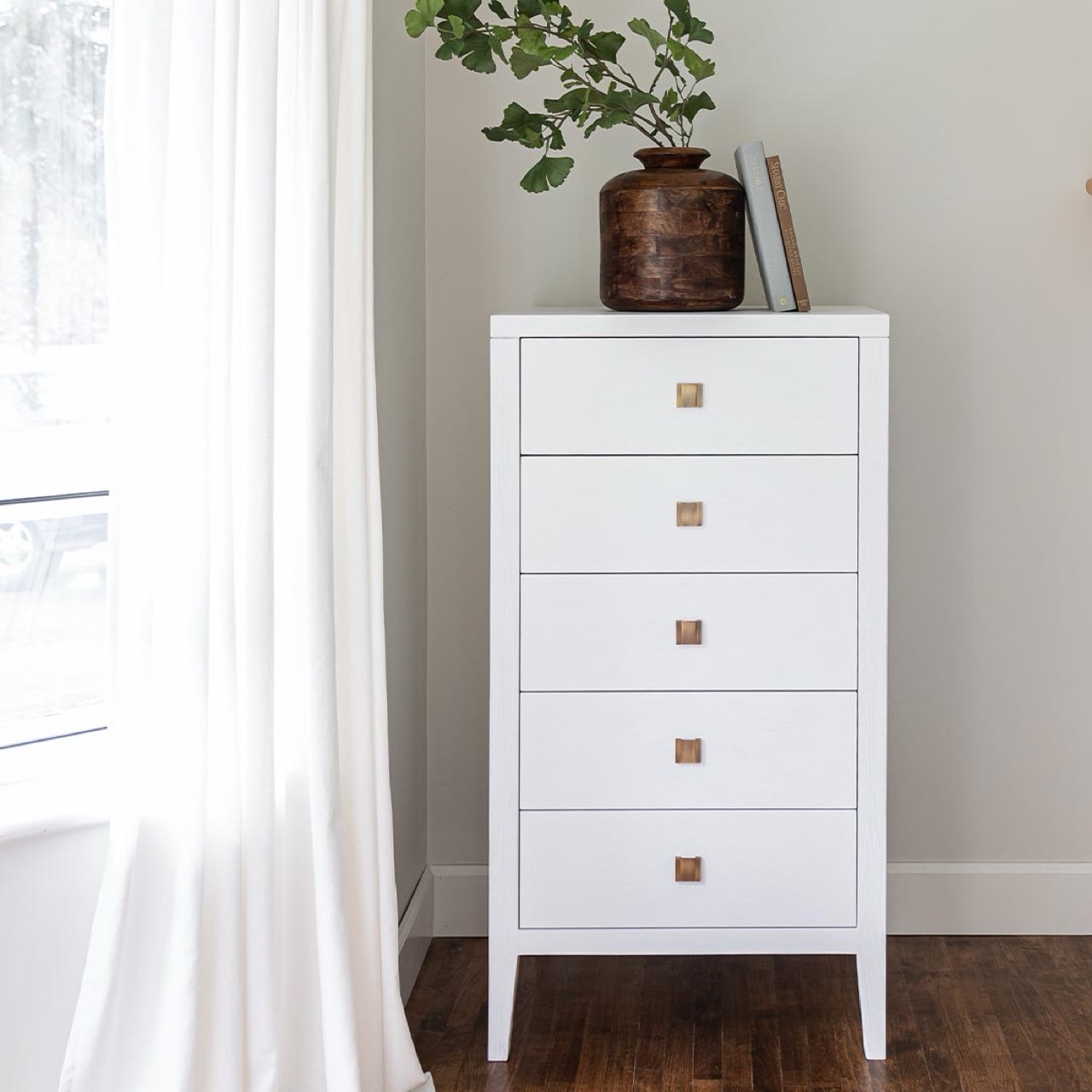 Studio lifestyle shot featuring the white tall dresser styled with a decorative vase in a bright, airy bedroom corner