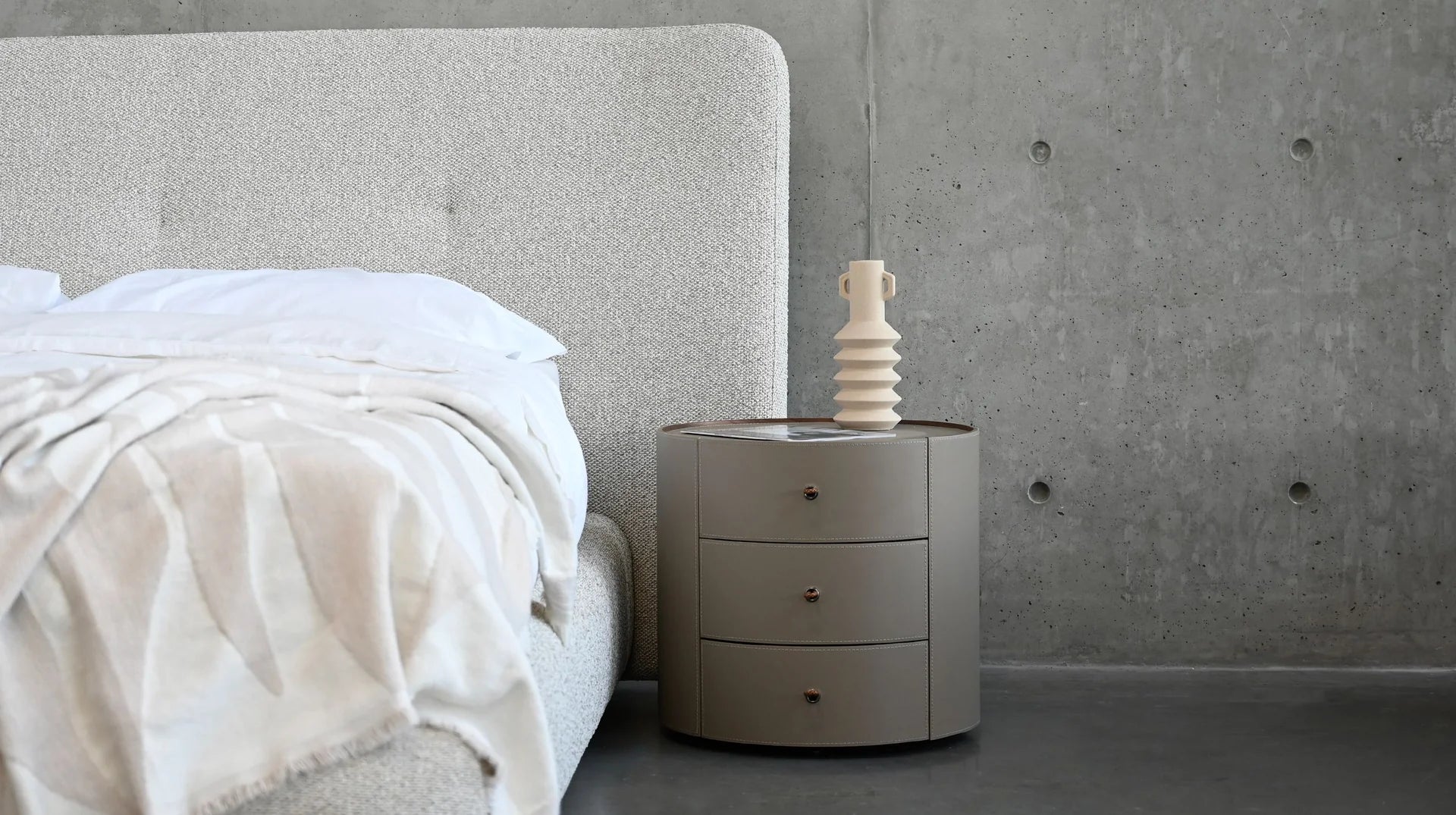 Front view of the Neru nightstand featuring a circular 3-drawer silhouette wrapped in light grey leather with contrasting top stitching