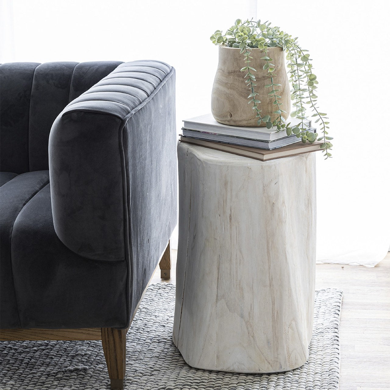 Lifestyle view of the 21-inch white wood stump used as a side table next to a dark grey upholstered sofa with books and plants