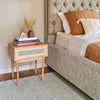 Lifestyle view of the natural cane side table used as a nightstand next to a tufted upholstered bed with warm autumn-toned bedding