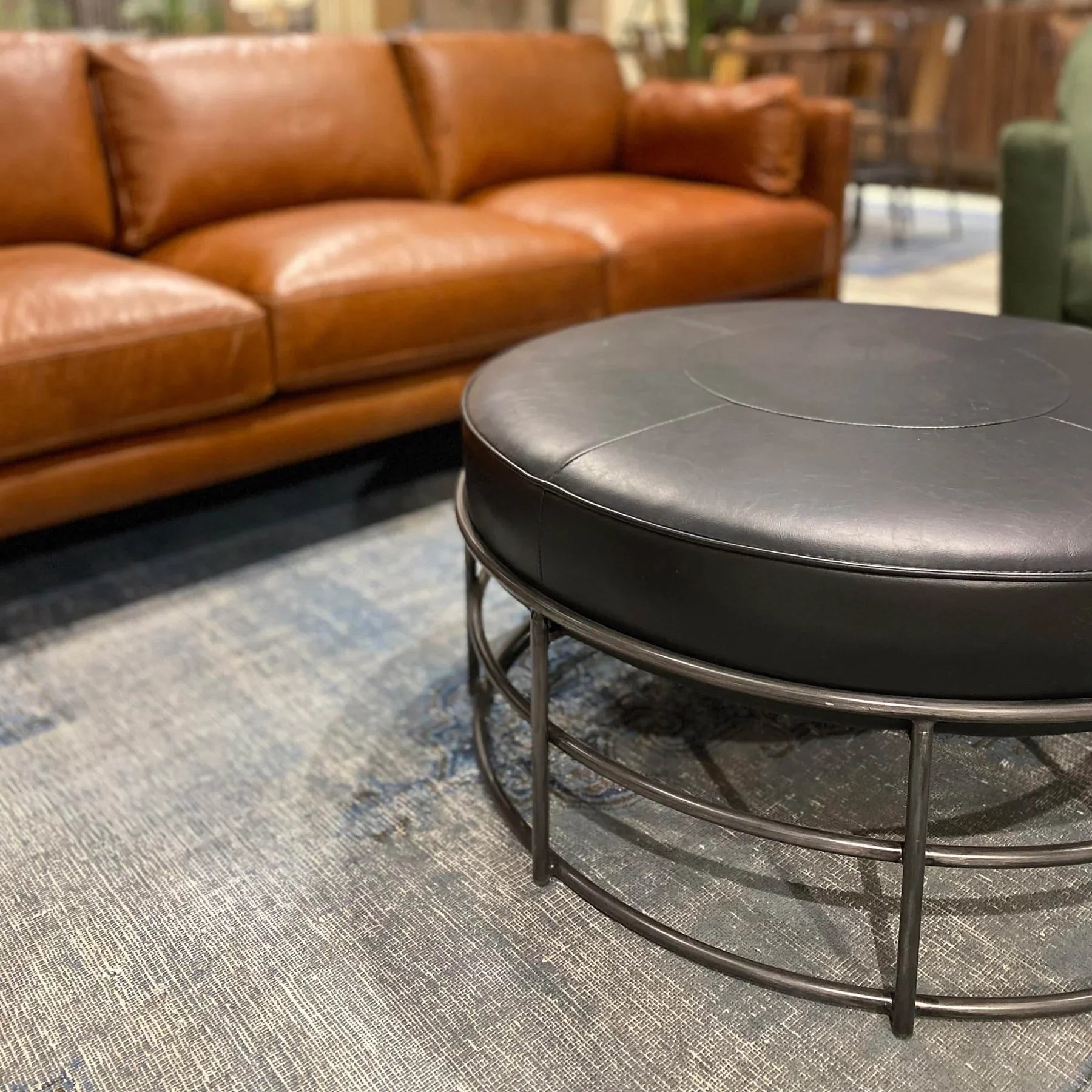 Front three-quarter view of the Ziggy round ottoman featuring a black leather top and a minimalist circular metal cage base